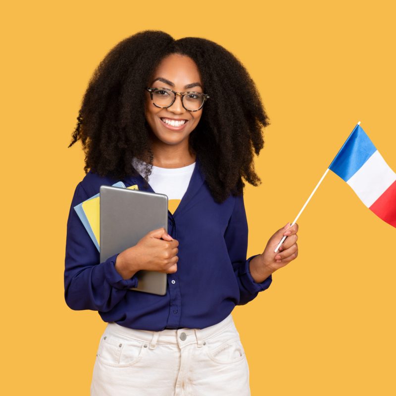 Excited african american lady student, with tablet and copybooks, holds flag of France and smiling on yellow background, enjoying foreign language education